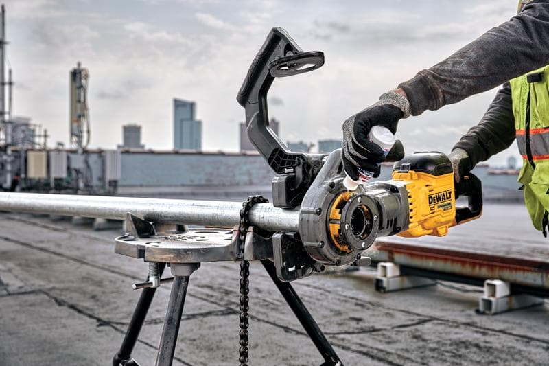 Uma pessoa usando uma ferramenta elétrica amarela DEWALT DCE700X2 para roscar um tubo de metal montado em um suporte de trabalho, ao ar livre em um telhado com edifícios da cidade visíveis ao fundo. O usuário está usando luvas e colete de segurança.