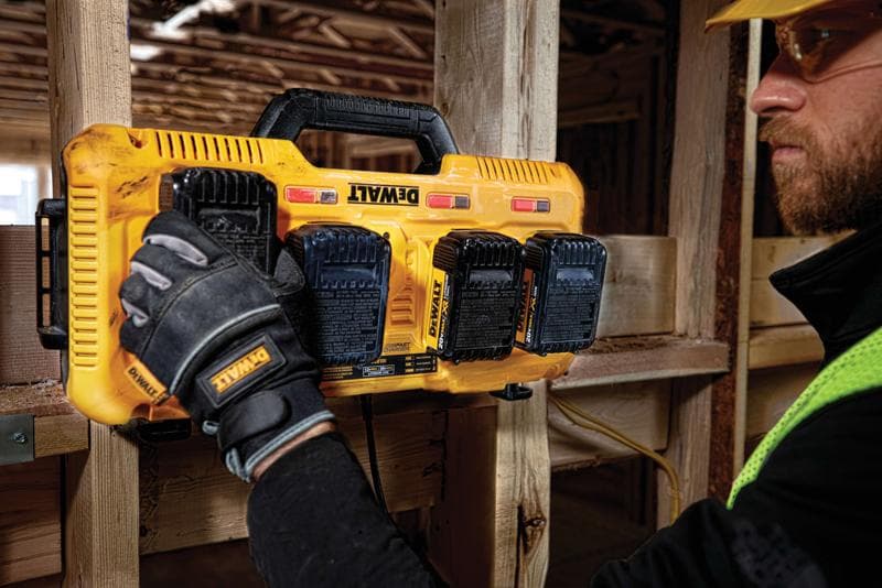 Uma pessoa usando luvas de trabalho instala ou remove baterias de um carregador múltiplo amarelo DEWALT montado em uma viga de madeira em um canteiro de obras.