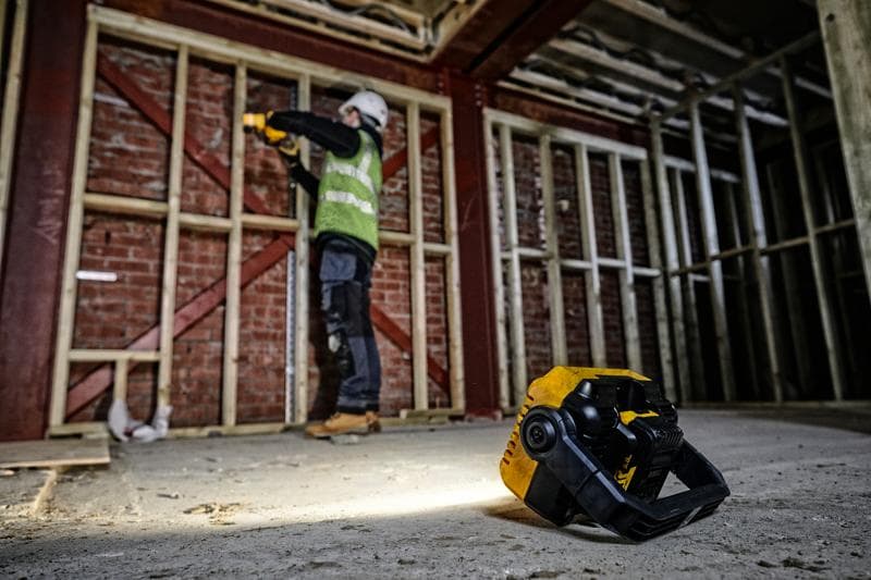 Luz de trabalho DEWALT DCL077 no chão, trabalhador ao fundo junto a estrutura de madeira.