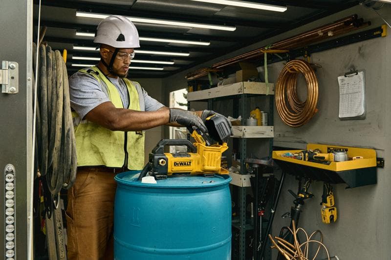 Uma pessoa usando capacete de segurança e colete está utilizando uma ferramenta DEWALT, modelo DCE050P1, em cima de um barril azul dentro de um espaço de oficina ou armazenamento. O local possui prateleiras, tubos de cobre enrolados e outras ferramentas e equipamentos visíveis.