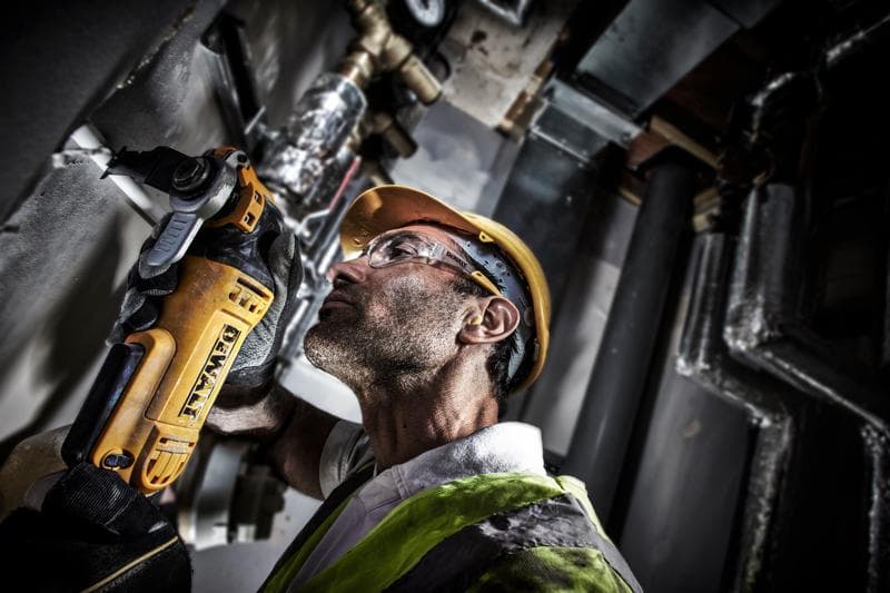 Trabalhador usa DEWALT DWE315 para cortar tubo, com capacete e colete em ambiente industrial.