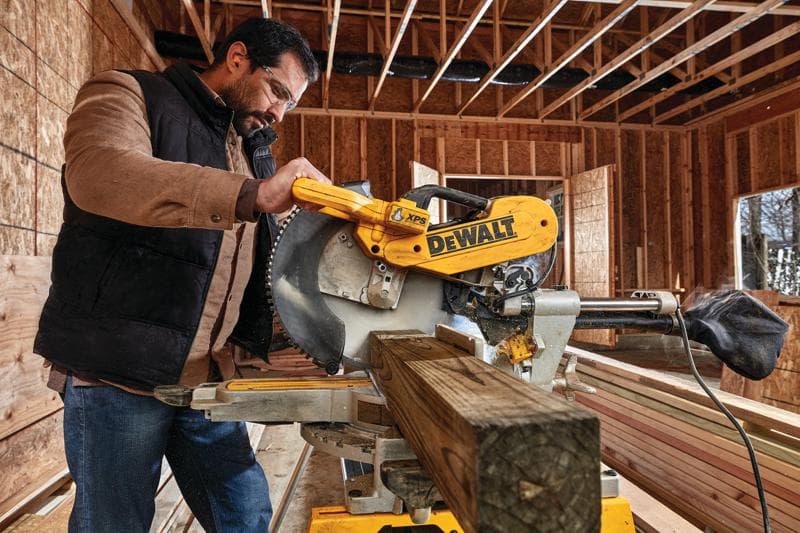 Uma pessoa opera uma serra de esquadria DEWALT DWS780 para cortar uma viga de madeira em um local de construção. A serra é amarela e exibe o logotipo da DEWALT em destaque. O fundo mostra estruturas de madeira expostas e paredes inacabadas.