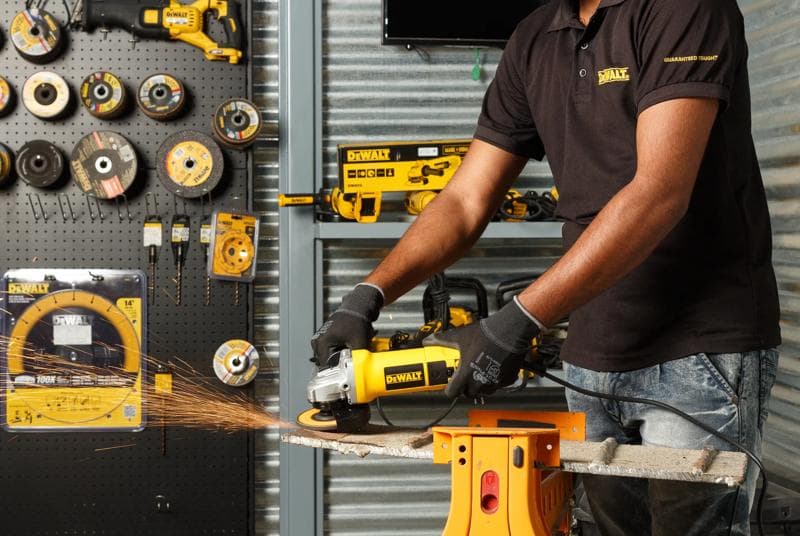 Uma pessoa usando luvas opera uma esmerilhadeira angular Dewalt DW803 para cortar uma peça de metal, com faíscas voando. Vários ferramentas elétricas Dewalt e discos de esmerilhagem estão exibidos em uma parede e bancada ao fundo.