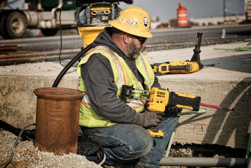 Um operário da construção usando capacete amarelo, colete de segurança e luvas está utilizando uma ferramenta elétrica DEWALT para perfurar concreto em um canteiro de obras ao ar livre. O rosto do trabalhador está borrado para preservar a privacidade. Outras ferramentas DEWALT e equipamentos de construção aparecem ao fundo.
