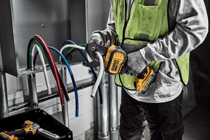 Uma pessoa usando luvas e colete de segurança está utilizando um cortador de cabos sem fio DEWALT para cortar um cabo elétrico branco grosso. Vários cabos coloridos estão visíveis saindo de um painel elétrico.