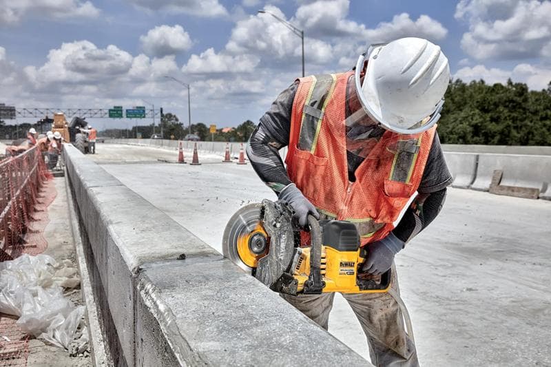 Um trabalhador da construção civil usando capacete branco e colete de segurança laranja está utilizando uma serra sem fio DEWALT (modelo DCS692B) para cortar uma barreira de concreto em um canteiro de obras.