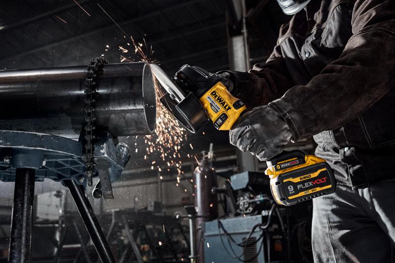 Uma pessoa usando equipamento de proteção utiliza uma esmerilhadeira angular sem fio DEWALT para cortar um grande tubo de metal em uma oficina industrial, com faíscas voando durante o corte.