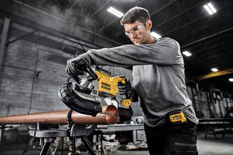 Uma pessoa usa uma serra de fita portátil DEWALT DCS374B para cortar um tubo de metal em uma oficina industrial. O rosto da pessoa está desfocado para preservar a privacidade.